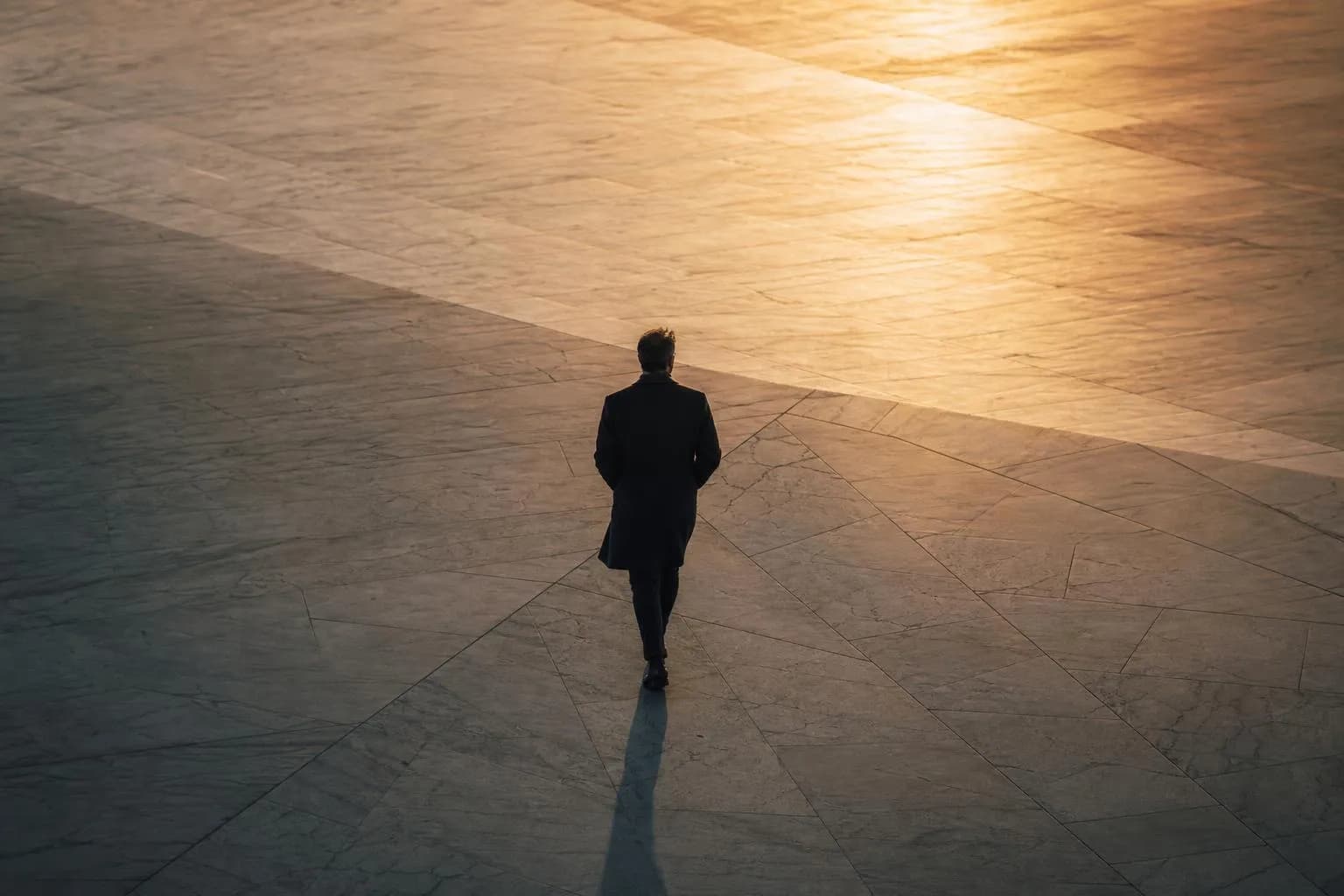 Solitary figure walking across expansive plaza at dusk, gold light, minimal composition