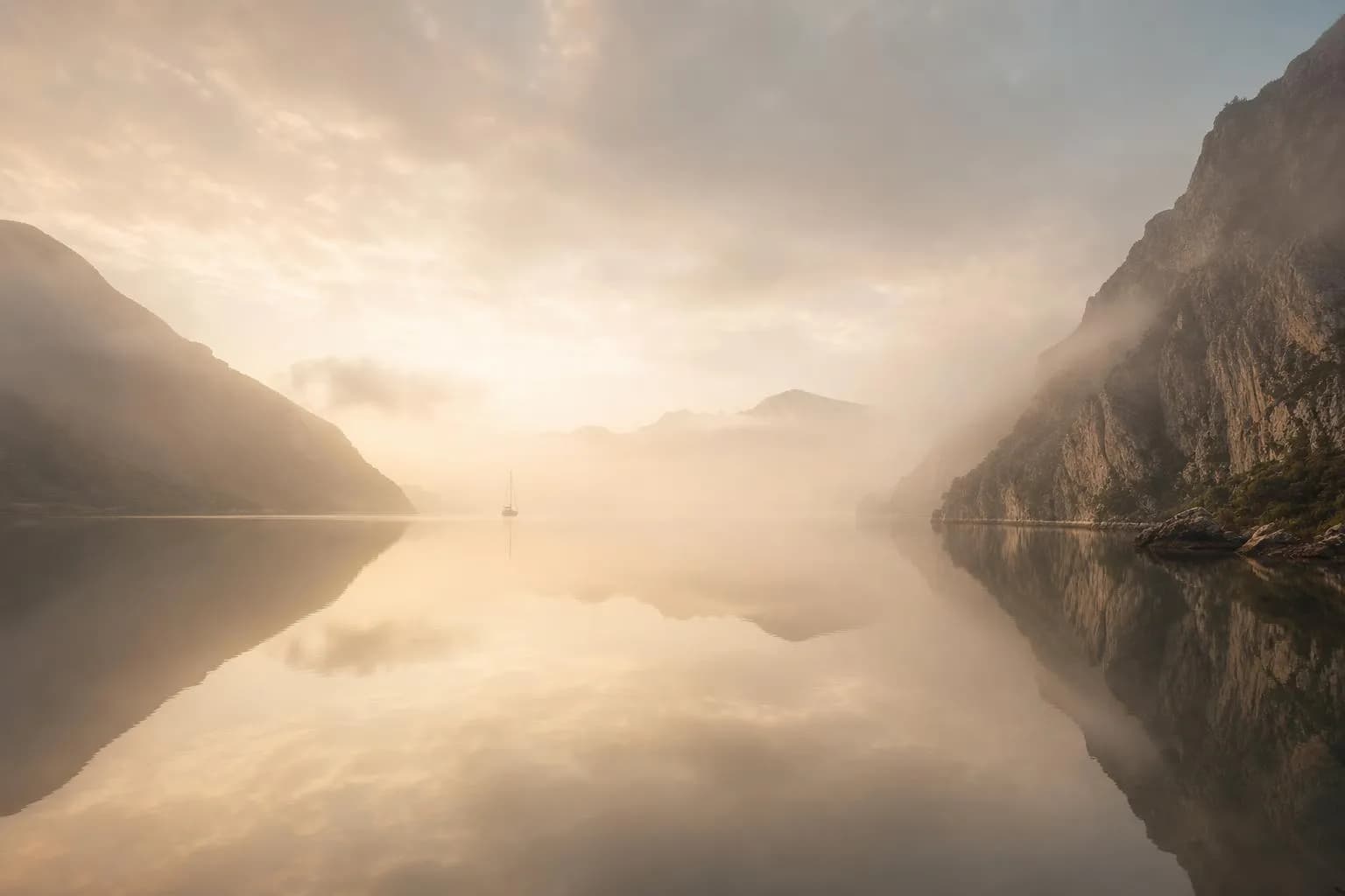 Misty coastal cove at dawn with still water and distant anchored sailboat.