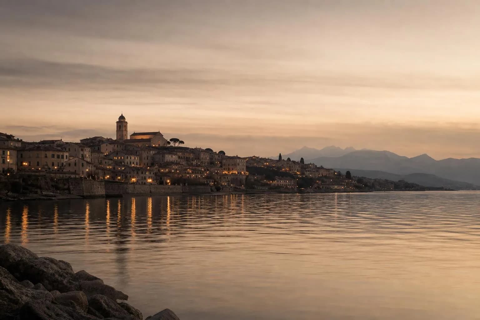 Distant European coastal town at dusk, viewed across calm water under a pale sky
