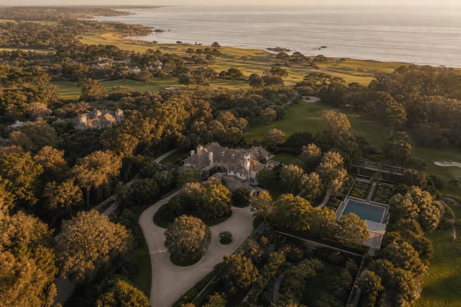 Aerial view of a manicured private estate with winding paths and distant coastline at golden hour.