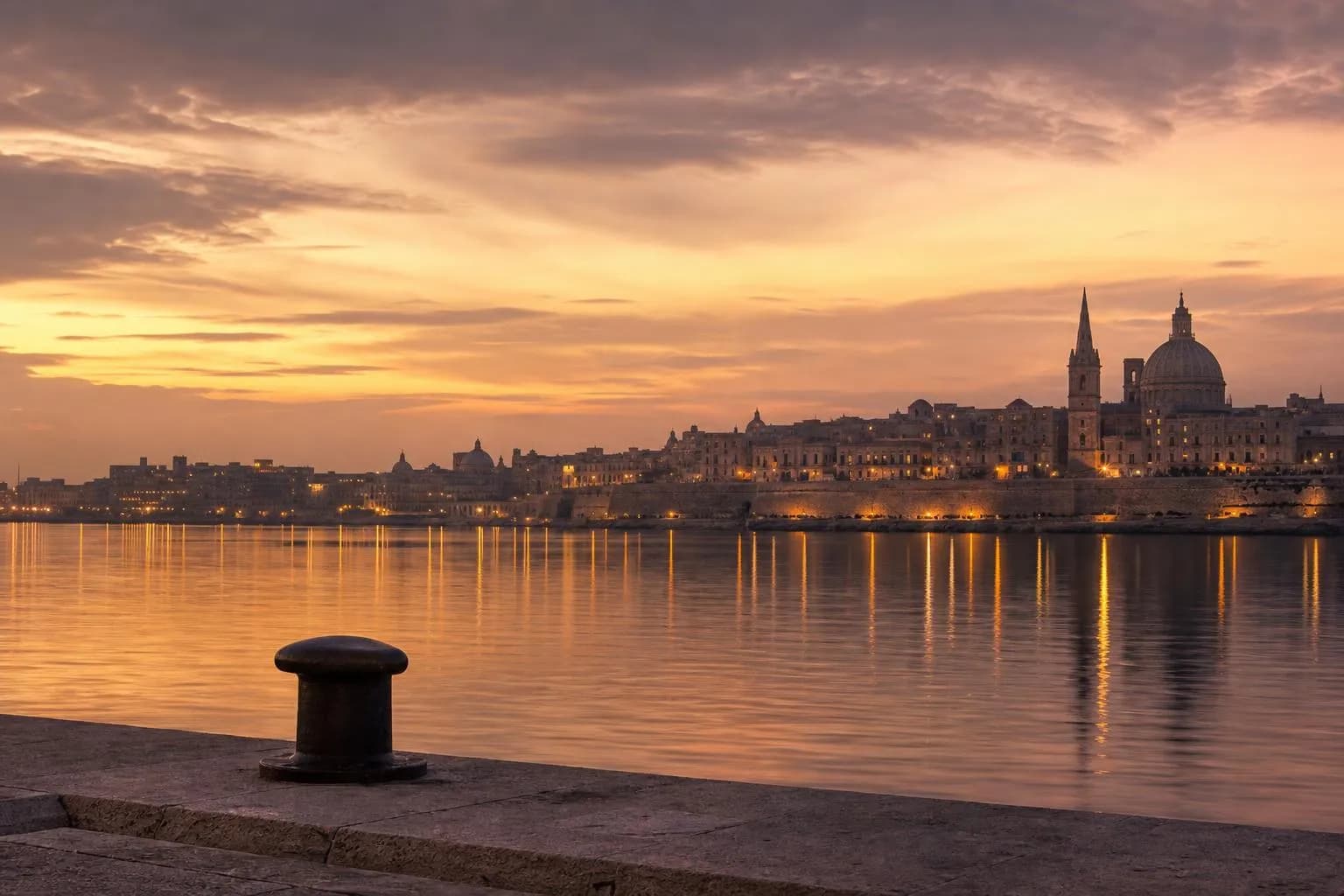 Distant cityscape reflected in calm water at dusk, viewed from a quiet shoreline