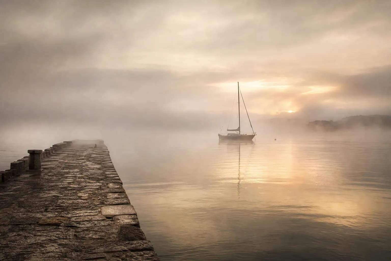 Misty harbor at dawn with a lone sailboat anchored in still water and a weathered pier.