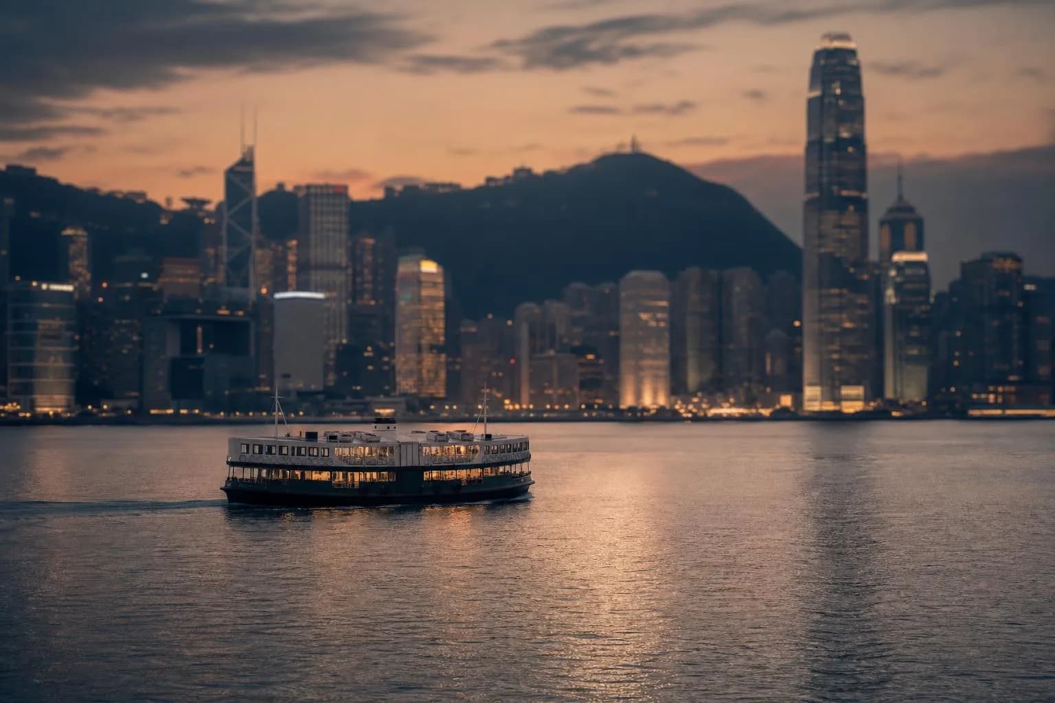 Hong Kong's Victoria Harbour at dusk, with a ferry crossing calm water and the city skyline lit softly beyond.