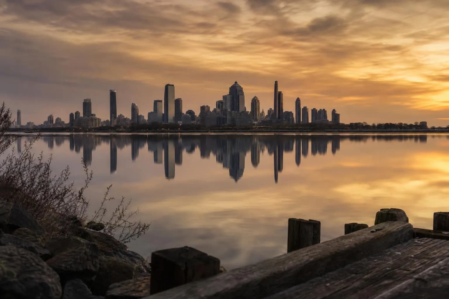 Distant city skyline reflected in still water at dusk, viewed from quiet shoreline.