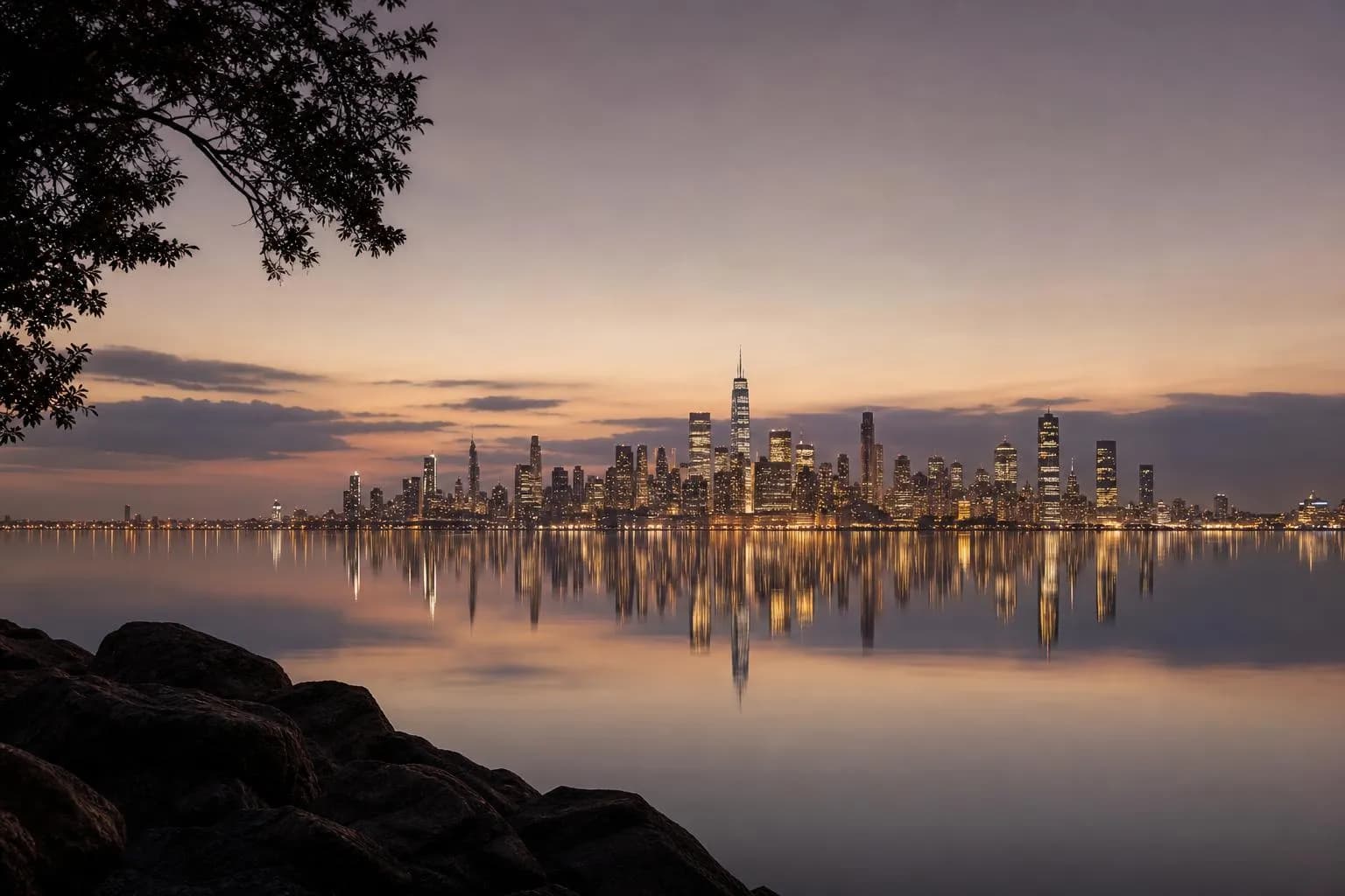 Distant city skyline glowing at dusk across calm water, soft lavender sky, quiet and reflective.