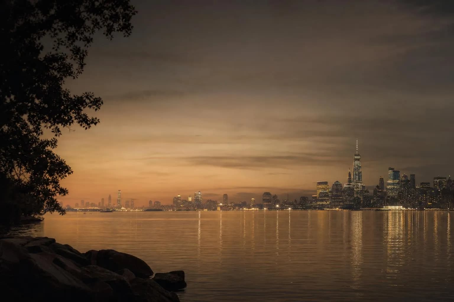 Distant city skyline glowing at dusk across calm water, viewed from a peaceful shoreline.