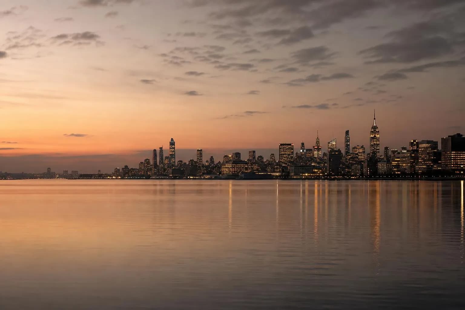 Distant city skyline reflected in calm dark water at dusk, viewed from afar across still water.