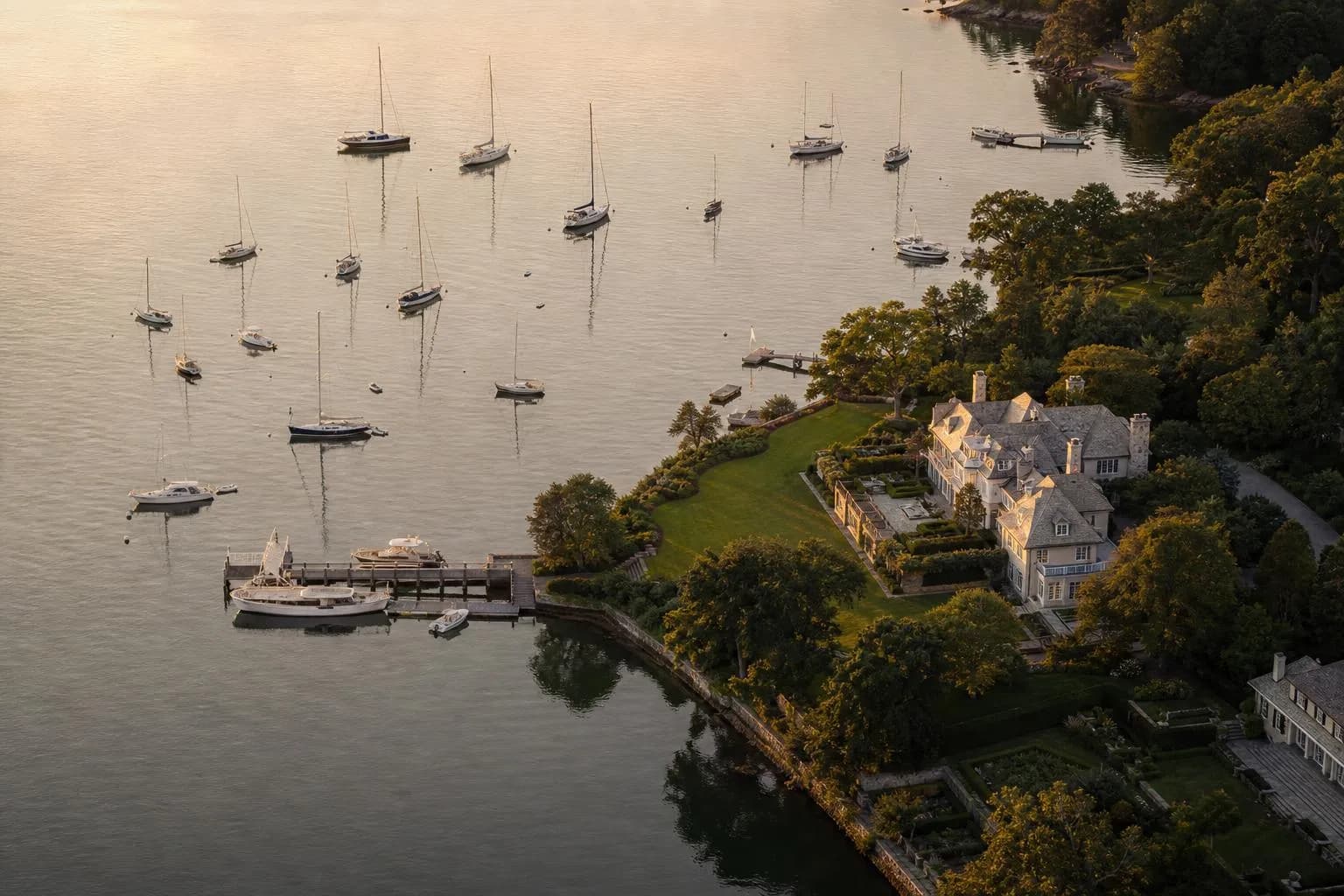 Aerial view of a serene harbor with moored boats and a waterfront estate at golden hour.