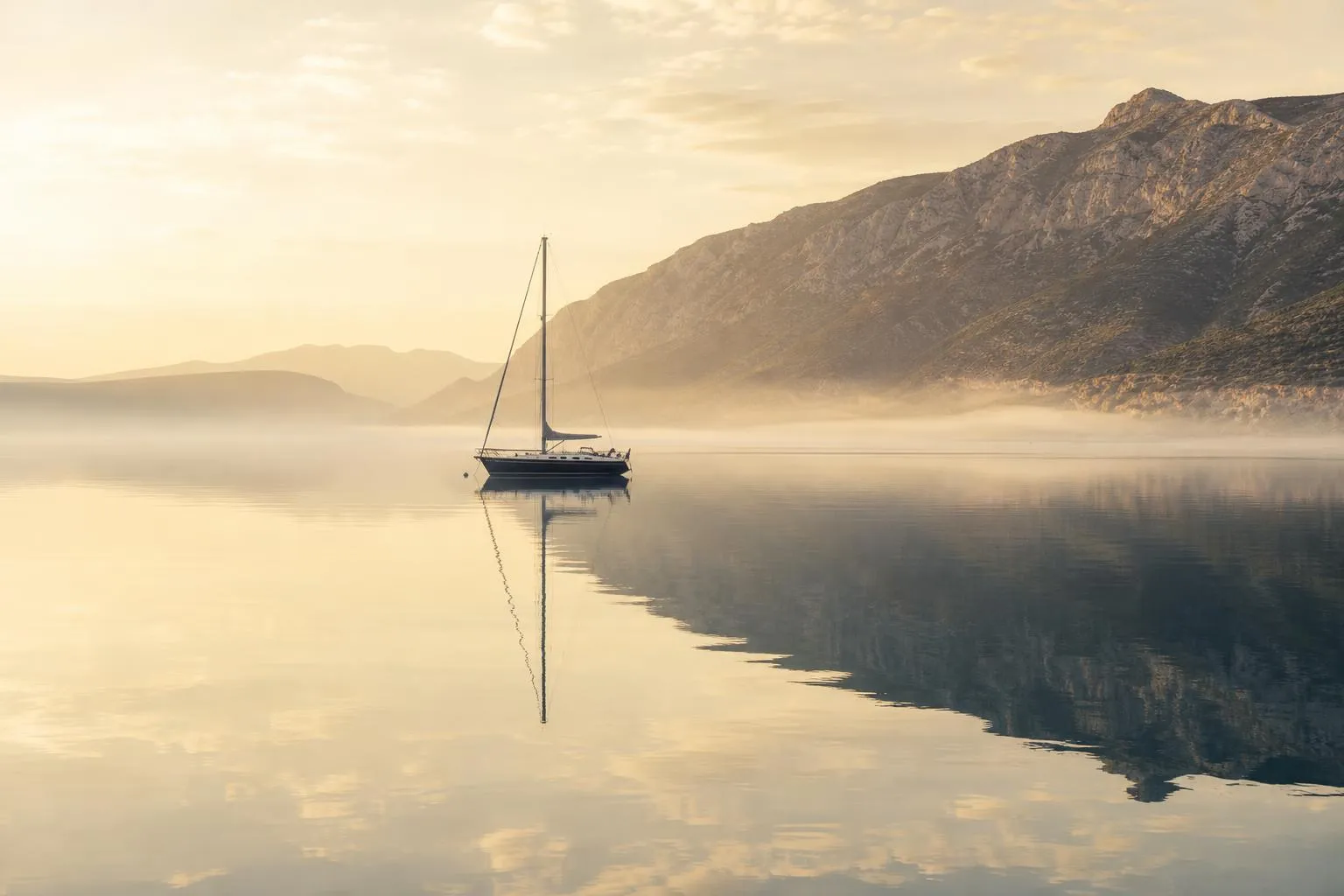 Serene harbor at dawn with lone sailboat, still water, and misty cliffs—calm, undisturbed landscape