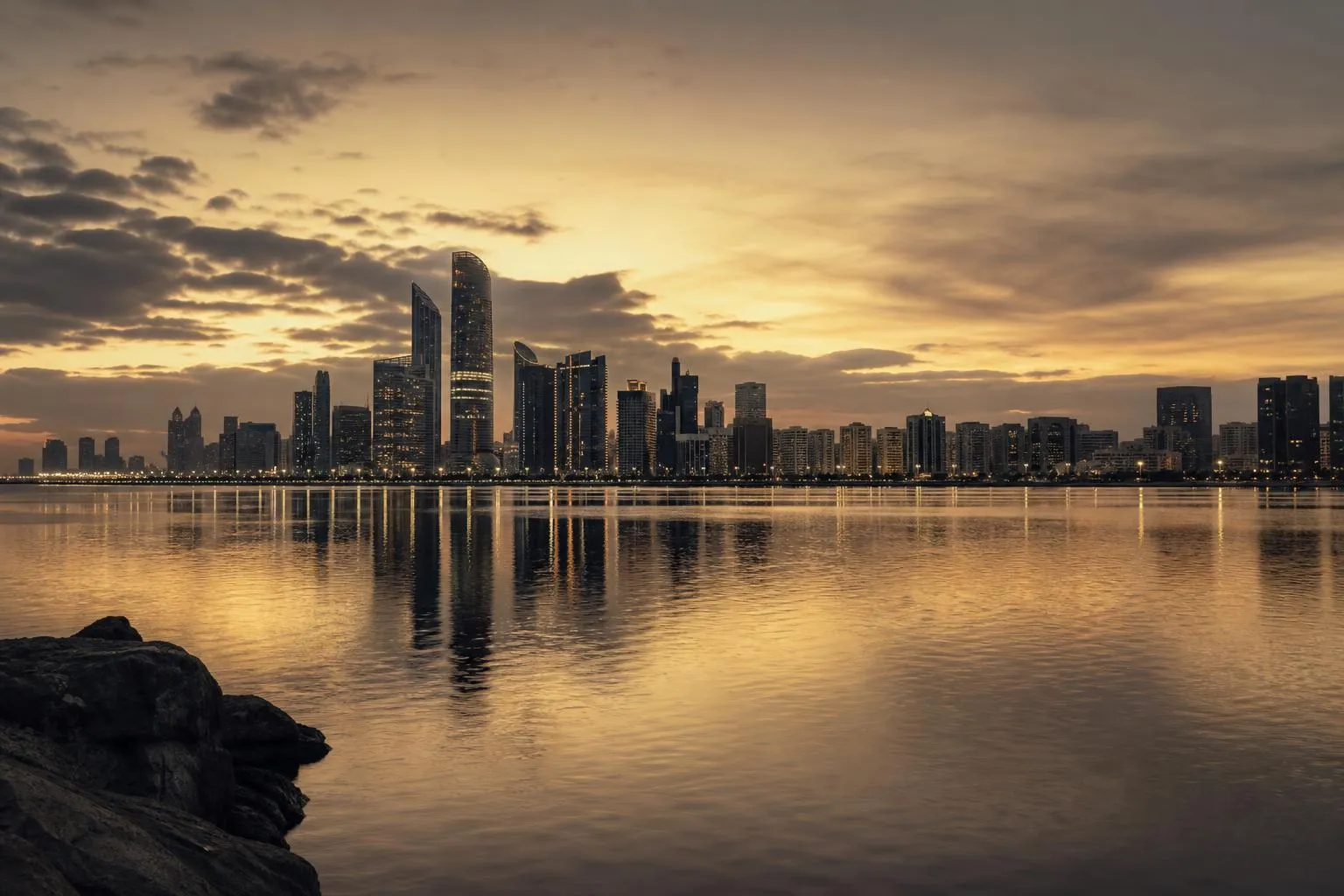 Distant city skyline glowing at dusk, reflected in still water, viewed from a quiet shoreline.