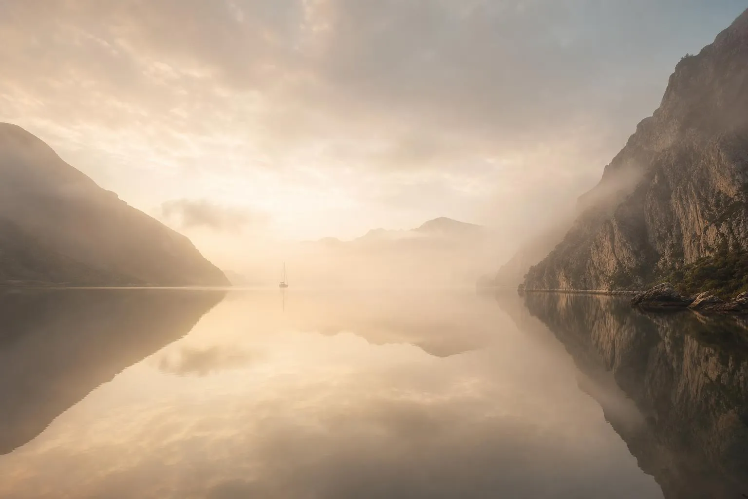 Misty coastal cove at dawn with still water and distant anchored sailboat.