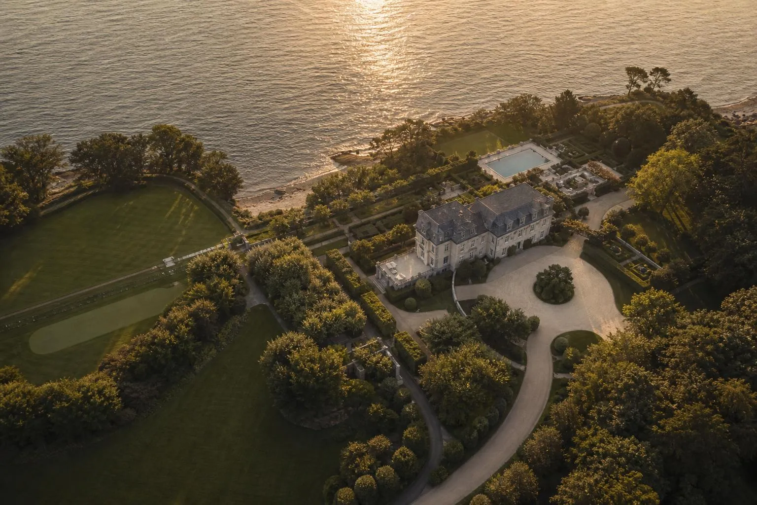 Aerial view of a coastal estate with manicured grounds and private drive at sunset.