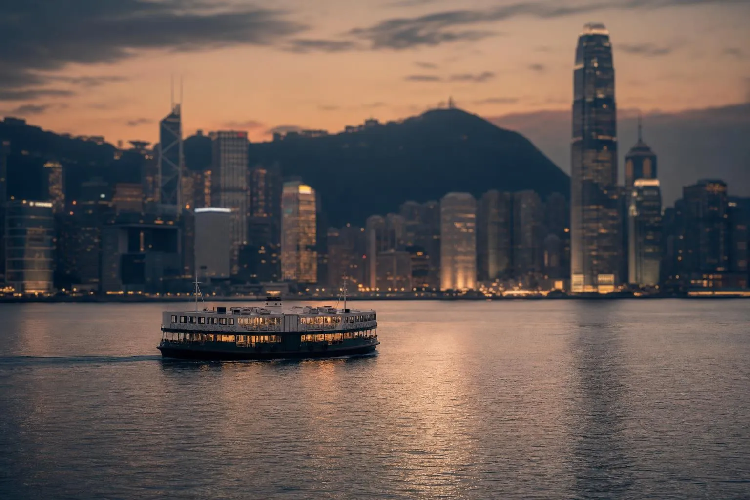 Hong Kong's Victoria Harbour at dusk, with a ferry crossing calm water and the city skyline lit softly beyond.