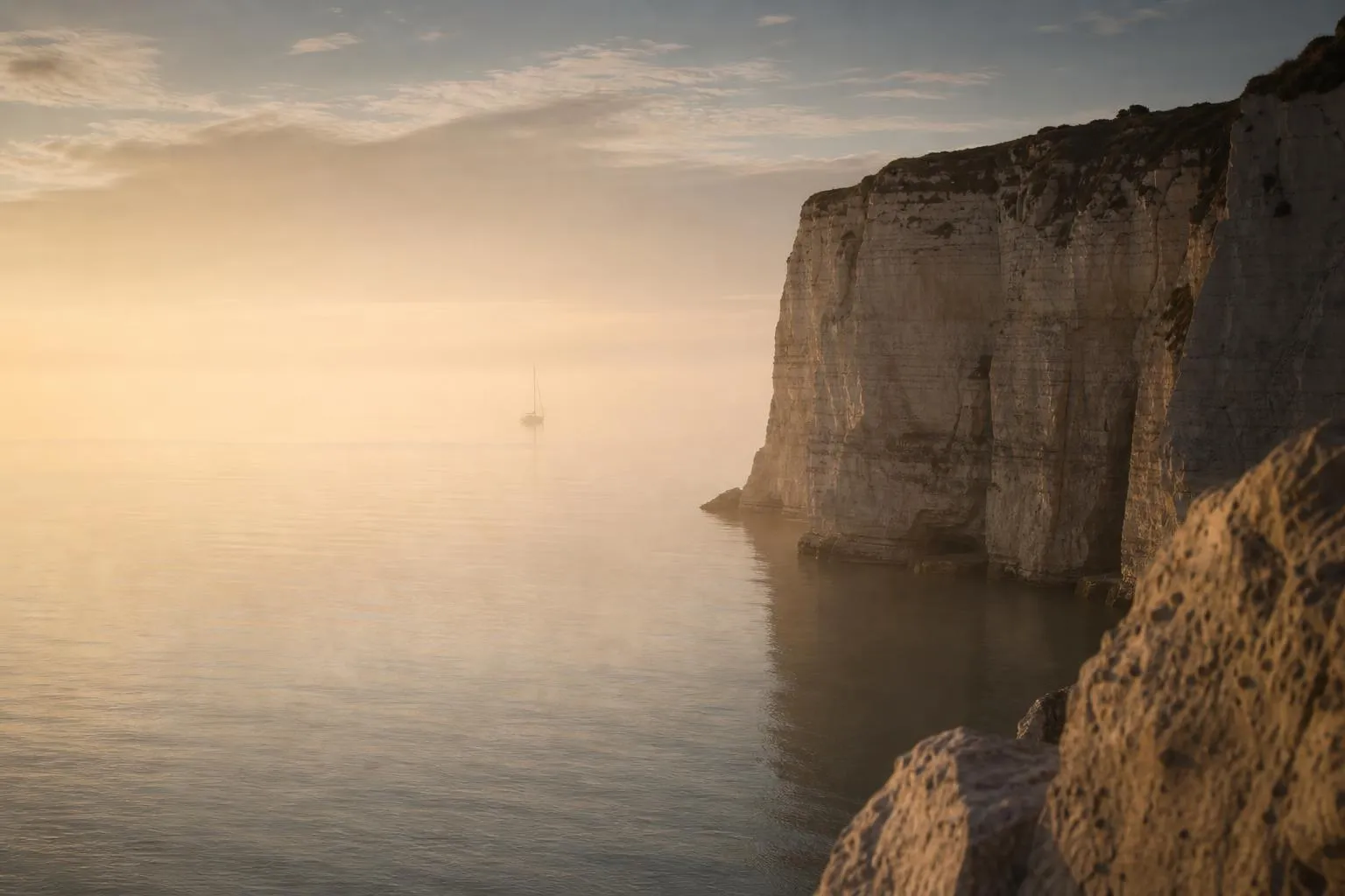Misty coastal cliffs and still water at dawn, lone sailboat on horizon.
