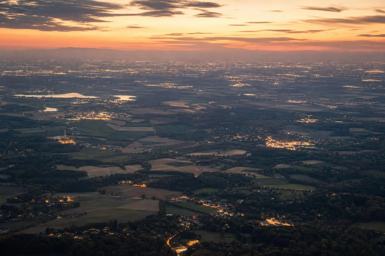Aerial view of varied landscape at dusk, distant city lights glowing softly across the horizon